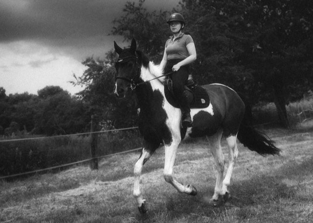 Black-and-white photograph of a rider on a piebald horse moving across a grassy field beneath dramatic clouds.